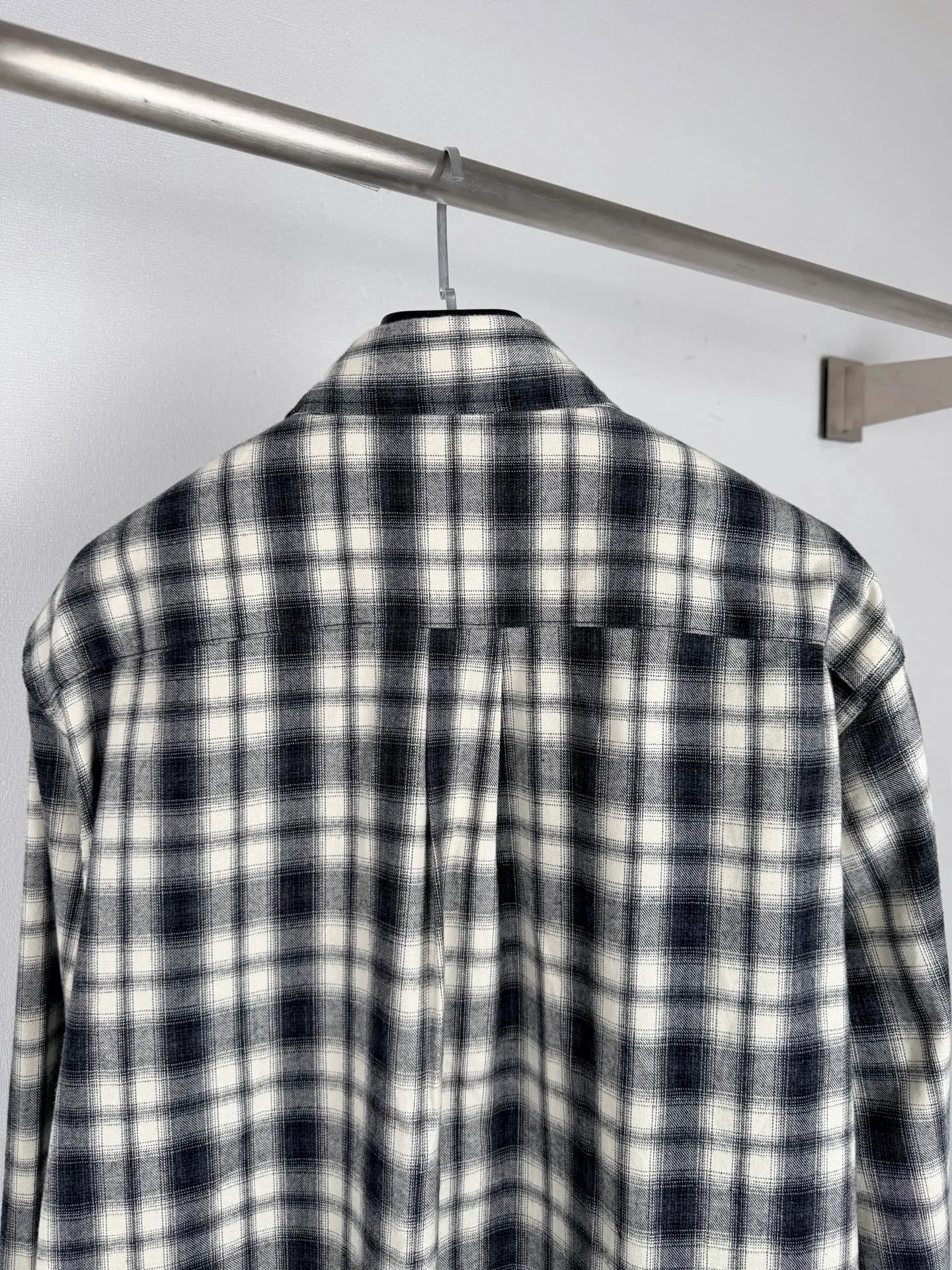 BALENCIAGA/balenciaga Black White plaid pocket embroidered logo shirt jacket The technical difficulties of this plaid shirt focus on fabric finishing, plaid matching and component sewing details. The fabric has been sanded and distressed to create a fine velvet feel on the surface, changing the original luster of the fabric and making the Black and White plaid colors transition softly. At the same time, the drape properties of the fabric itself are retained, taking into account the looseness of the upper body and the silhouette of the body, without appearing soft and shapeless. The biggest test of plaid garments comes from the alignment of the pieces. The front and rear body, large and small sleeves, and chest pockets are all independent pieces. In the cutting stage, the horizontal and vertical axes of the plaid need to be aligned, and the external chest bag is cut separately. The plaid on the bag surface needs to form a visual coordination with the main body of the garment. Cutting and typesetting will produce additional fabric loss, and there are high requirements for the cutting accuracy of the cutting bed typesetting and workers. The collar adopts a double-layer combined structure, and the collar surface and collar seat are double-stitched with even and regular stitching spacing to ensure the lapel's long-lasting shape and resistance to warping and deformation after repeated bending. The placket is sewn with regular topstitching and paired with Black matte buttons. The button points need to be aligned with the checkered lines to avoid damaging the overall visual order. The splicing of the cuffs also takes into account the grain direction, and the overlocking process reinforces the seams to improve the durability of the garment. After the entire garment is sewn, it also undergoes a garment washing process to further strengthen the Retro texture of the fabric, unify the tone and texture of the entire garment, weaken the harshness of industrial production, and finally present a relaxed Retro garment effect. Size: 46 48 50 52